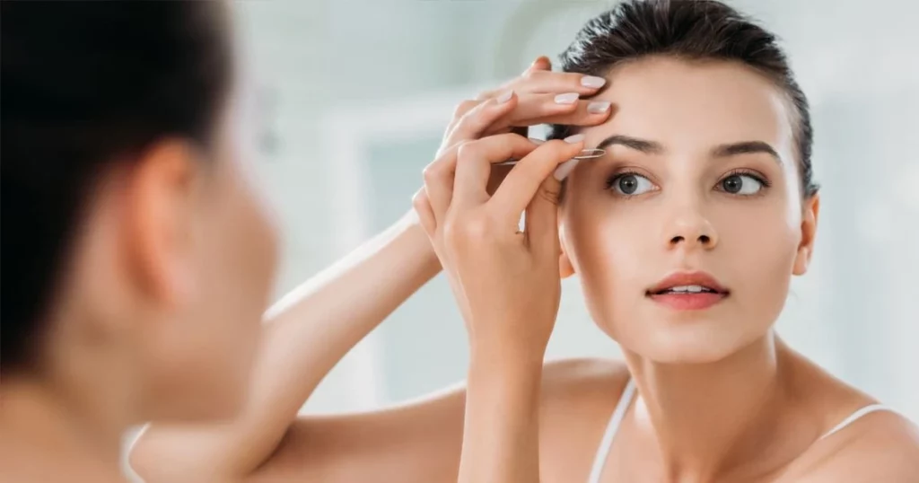 Une femme qui se regarde dans le miroir et qui s'épile les sourcils avec une pince à épiler